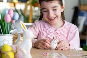 A cute girl with pink bunny ears makes an Easter craft - decorates an egg in the form of a unicorn with rhinestones, horn, flowers in the interior of a house with plants. photo