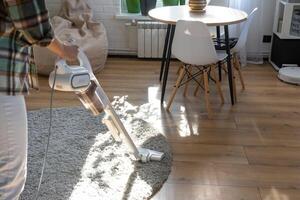 A woman vacuums a round carpet in a house among house plants with a hand vacuum cleaner. General cleaning of the house, cleaning service and housewife photo