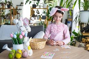 A cute girl with pink bunny ears makes an Easter craft - decorates an egg in the form of a unicorn with rhinestones, horn, flowers in the interior of a house with plants. photo