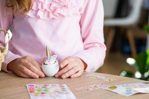 A cute girl with pink bunny ears makes an Easter craft - decorates an egg in the form of a unicorn with rhinestones, horn, flowers in the interior of a house with plants. photo