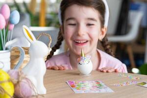 A cute girl with pink bunny ears makes an Easter craft - decorates an egg in the form of a unicorn with rhinestones, horn, flowers in the interior of a house with plants. photo