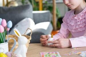 A cute girl with pink bunny ears makes an Easter craft - decorates an egg in the form of a unicorn with rhinestones, horn, flowers in the interior of a house with plants. photo