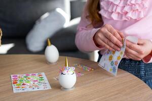 A cute girl with pink bunny ears makes an Easter craft - decorates an egg in the form of a unicorn with rhinestones, horn, flowers in the interior of a house with plants. photo