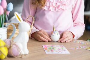 un linda niña con rosado conejito orejas hace un Pascua de Resurrección arte - decora un huevo en el formar de un unicornio con pedrería, bocina, flores en el interior de un casa con plantas. foto