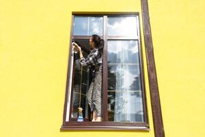 un mujer a mano lavados el ventana de el casa con un trapo con un rociar limpiador y un fregona afuera. la seguridad a altura, restaurar orden y limpieza en el primavera, limpieza Servicio foto