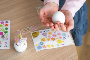 A cute girl with pink bunny ears makes an Easter craft - decorates an egg in the form of a unicorn with rhinestones, horn, flowers in the interior of a house with plants. photo