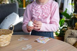 A cute girl with pink bunny ears makes an Easter craft - decorates an egg in the form of a unicorn with rhinestones, horn, flowers in the interior of a house with plants. photo