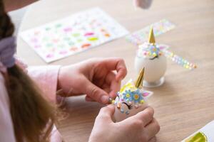 A cute girl with pink bunny ears makes an Easter craft - decorates an egg in the form of a unicorn with rhinestones, horn, flowers in the interior of a house with plants. photo