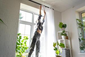 Woman hangs transparent tulle curtains on large windows in the house inside the interior. Spring cleaning, tidying up. photo