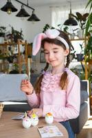 A cute girl with pink bunny ears makes an Easter craft - decorates an egg in the form of a unicorn with rhinestones, horn, flowers in the interior of a house with plants. photo