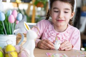 un linda niña con rosado conejito orejas hace un Pascua de Resurrección arte - decora un huevo en el formar de un unicornio con pedrería, bocina, flores en el interior de un casa con plantas. foto