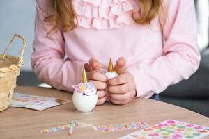 un linda niña con rosado conejito orejas hace un Pascua de Resurrección arte - decora un huevo en el formar de un unicornio con pedrería, bocina, flores en el interior de un casa con plantas. foto