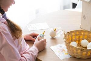 A cute girl with pink bunny ears makes an Easter craft - decorates an egg in the form of a unicorn with rhinestones, horn, flowers in the interior of a house with plants. photo