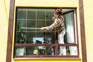 un mujer a mano lavados el ventana de el casa con un trapo con un rociar limpiador y un fregona afuera. la seguridad a altura, restaurar orden y limpieza en el primavera, limpieza Servicio foto