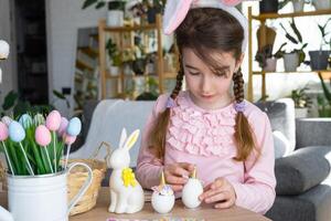 un linda niña con rosado conejito orejas hace un Pascua de Resurrección arte - decora un huevo en el formar de un unicornio con pedrería, bocina, flores en el interior de un casa con plantas. foto