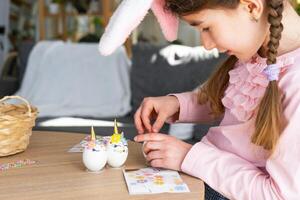 A cute girl with pink bunny ears makes an Easter craft - decorates an egg in the form of a unicorn with rhinestones, horn, flowers in the interior of a house with plants. photo