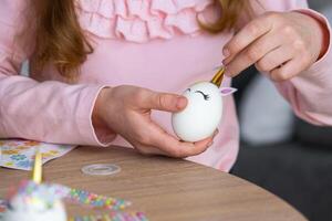 A cute girl with pink bunny ears makes an Easter craft - decorates an egg in the form of a unicorn with rhinestones, horn, flowers in the interior of a house with plants. photo