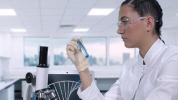 A young female laboratory technician in a white coat conducts laboratory tests of a blue fluid in a test tube while sitting at a table in a science lab. Close-up plan.slow motion video