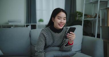 métrage lent mouvement tir, content Jeune asiatique femme asseoir sur canapé en portant café tasse et prendre plaisir prise selfies sur mobile téléphone à à la maison, gratuitement le temps, prends pause, sourire video