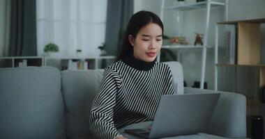 beeldmateriaal dolly geschoten, blij jong Aziatisch vrouw zitten Aan sofa drinken koffie of thee terwijl werken creatief baan van huis Aan laptop met online netwerk, nemen pauze, glimlachend video