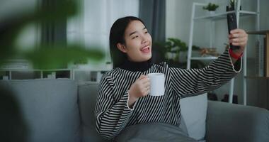 Footage dolly shot, Happy young asian woman sit on sofa holding coffee cup and enjoy taking selfies on mobile phone at home,Free time,take break,smiling video