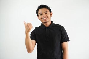 smiling young asian man against a vibrant white studio background, thumb pointing to the right side, with thumb finger away, laughing and carefree, wearing black polo t shirt. photo