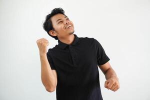 sonriente o contento joven asiático hombre apretado su puños aplausos despreocupado y entusiasmado. victoria concepto, vistiendo negro polo t camisa aislado en blanco antecedentes foto