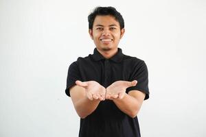 sonriente joven asiático hombre posando en un blanco fondo participación alguna cosa con palmas, ofrecimiento a cámara, vistiendo negro polo t camisa. foto