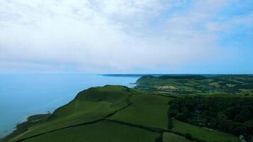 Aerial view of a lush green coastline with calm blue sea under a soft blue sky with light clouds. video