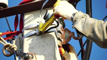 Skilled electrician in helmet fixes wires standing on ladder near high pole against blue sky on summer day backside view. Electrical service and mounting on the pole. Slow motion video