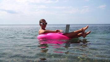 Woman freelancer works on laptop swimming in sea on pink inflatable ring. Happy tourist in sunglasses floating on inflatable donut and working on laptop computer in calm ocean. Remote working anywhere video