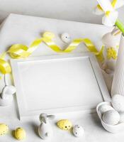 White table, square empty white frame, eggs, yellow ribbon, ceramic bunnies on white. Easter image. photo