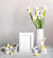 Easter white still life with blank frame, decorative eggs, bunnies, vase with Iris flowers on table. photo