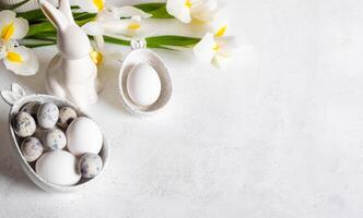 Easter white holiday card. Natural colors eggs in figured bowls, ceramic bunny, flowers. Copy space. photo