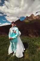 un joven hombre y su esposa estar en un abrazo alto en el montañas en contra el fondo de épico rocas en un soleado día. recién casados Boda Pareja en el montañas foto
