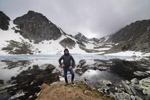 Happy active cheerful tourist in mountain sunglasses and a down jacket, joyful smiles expressively rejoices against the backdrop of a mountain lake high in the mountains photo