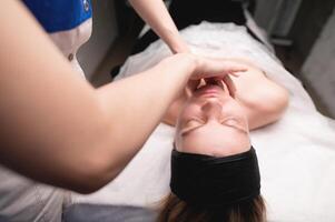 cosmético facial masaje procedimiento. de cerca de el cara de un joven mujer acostado en su atrás, haciendo un cara levantamiento masaje, ratería y laminación técnica foto