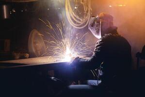 Young mechanic with a welding machine in an old dirty garage at night. Man in protection doing welding work, hobby photo