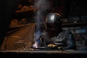 joven mecánico con un soldadura máquina en un antiguo sucio garaje a noche. hombre en proteccion haciendo soldadura trabajar, pasatiempo foto