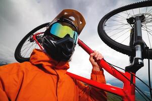 Wide angle vertical frame top angle. Portrait of a bearded mountain biker with his bicycle against a background of mountains and clouds in the summer surrounded by green grass photo