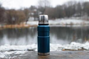 Vacuum travel thermos standing on wooden table outdoors. Steel flask on background winter lake, close-up. photo