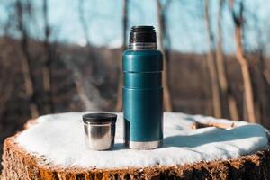 Thermos and mug with hot drink standing on stump. Winter forest, steam from warming drink in a cup. photo