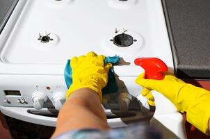 Female hand in rubber glove wiping the panel of gas stove, POV. Woman cleaning kitchen apartment with cleaning spray. Hygienic house cleaning photo