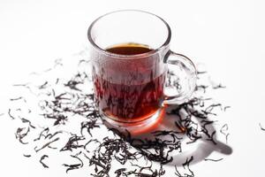 Glass cup of black tea and scattered dry ceylon tea leaves on white table, close up photo