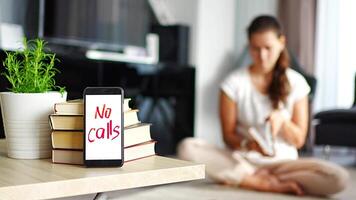 Digital detox concept photo. Smartphone with text No Calls and woman reading book in background. High quality 4k footage video