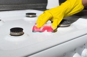 Female hand in glove cleaning the surface of gas stove with washing sponge and chemical foam. Home cleaning concept, kitchen hygiene photo