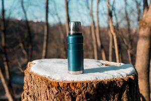 Thermos standing on tree stump covered with snow. Close-up of camping vacuum flask in the winter forest, sunny day. photo