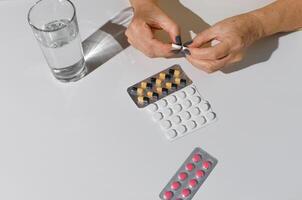 mayor mujer manos apertura paquete de medicamentos. tabletas y vaso de agua en blanco mesa, parte superior vista. foto
