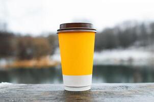 Cup of coffee standing on wooden table outdoors, close-up. Yellow paper glass on winter background. photo