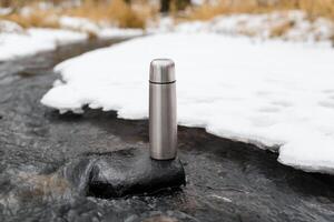 Steel vacuum thermos in a winter stream. Hiking flask standing on a stone outside. Adventure, hike, travel concept. photo